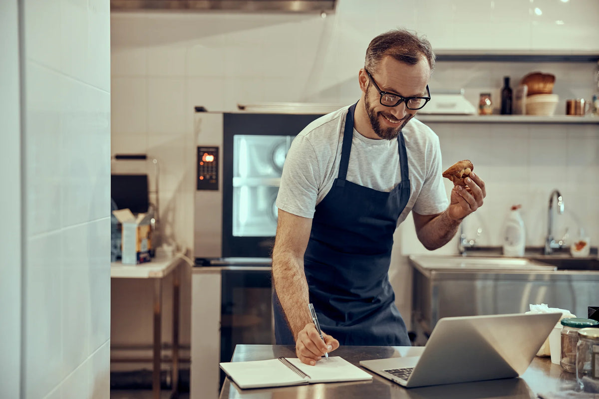 A man wearing a dark navy apron over a white t-shirt stands in a modern kitchen, writing in a notebook next to an open laptop while holding a piece of fried chicken
