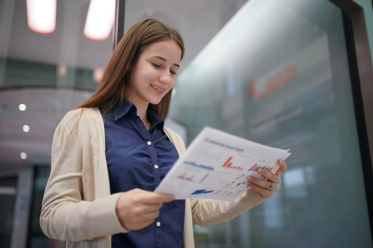 A woman in a beige cardigan and navy blue shirt looks at a printed chart or document in a modern office setting.